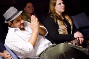 Man in fedora hat, hands together in praying formation looking up onto stage from the crowd