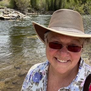 Susan Jane Gentile smiling, wearing a hat in from of a body of water.