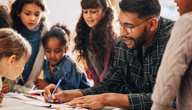 Teacher writing on table with several students surrounding and looking on.