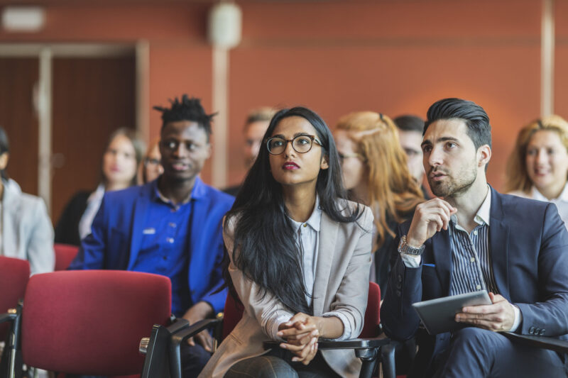 Business professionals seated in an auditorium, engaged with tablets during a presentation or meeting.