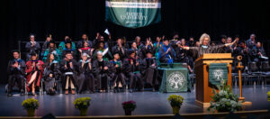 Stage with graduates and a speaker. Graduates are wearing a variety of regalia. There are green signs that say Antioch University and the speaker is at a podium. 