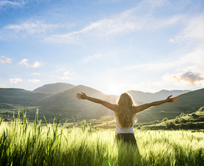 A woman stands in a vast field, arms outstretched, embracing the open space and the beauty of nature around her.