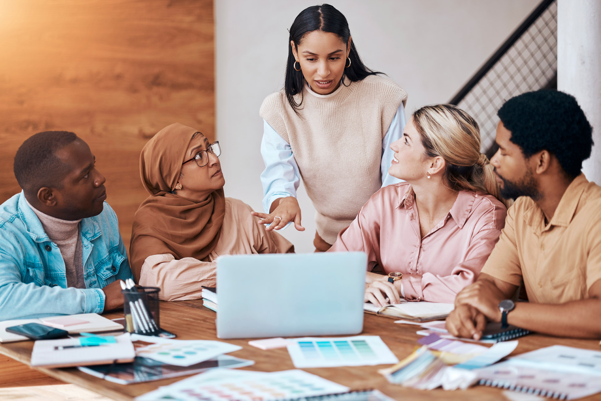 A group of diverse people working in an office, with a woman of color standing, leading a conversation
