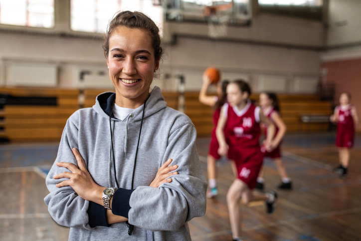 A woman in a sweatshirt stands confidently in a gym, arms crossed, showcasing a determined expression.